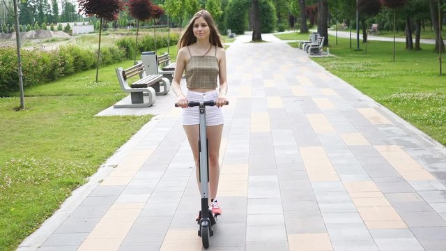 4K.Young long hair girl slowly  riding electric scooter in city park area. Steady shot, forward view