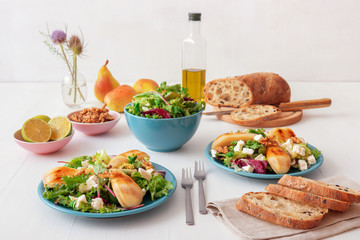 White wooden table with walnut salad, caramelised pears and feta cheese. Turquise dishes with salad, slices of twisted swiss bread with olives, limets, bottle of olive oil and whole pears.