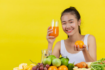 An Asian woman wearing a white tank top. Holding a glass of orange juice with your right hand The left hand held an orange and there were many fruits on the table.