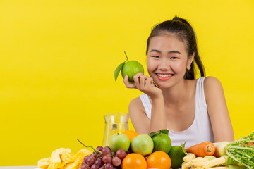 An Asian woman wearing a white tank top. Holding an orange with the right hand And on the table there are many different fruits.