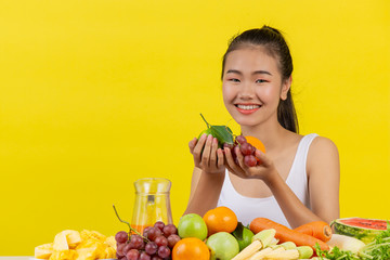 An Asian woman wearing a white tank top. Both hands held fruit and the table is full of various fruits.