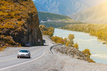 A winding asphalt black road with a yellow dividing strip between the rocks and the Katun River in the Altai mountains under a clear blue sky with clouds and a field covered with yellow autumn grass