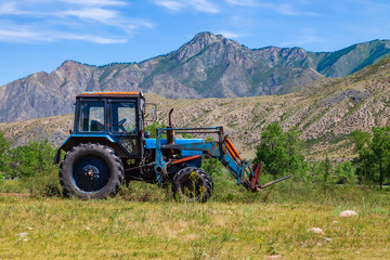 An old blue tractor excavator stands in a field with green and yellow grass against a background of mountains and blue sky during harvesting in summer or autumn. Agriculture and farming machinery.