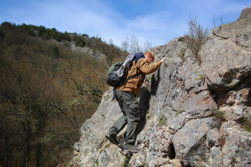Red-haired man in a brown windbreaker with a backpack and a rope climbs a cliff in the spring. In the background is a forest without foliage.