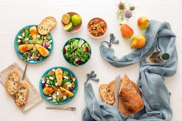 White wooden table with walnut salad, caramelised pears and feta cheese. Turquoise dishes with salad, slices of twisted swiss bread with olives, limets, bottle of olive oil and whole pears.