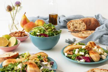 White wooden table with walnut salad, caramelised pears and feta cheese. Turquise dishes with salad, slices of twisted swiss bread with olives, limets, bottle of olive oil and whole pears.