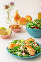 White wooden table with walnut salad, caramelised pears and feta cheese. Turquise dishes with salad, slices of twisted swiss bread with olives, limets, bottle of olive oil and whole pears.