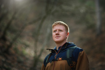 Handsome Caucasian man looks away and smiles slightly. Portrait of a red-haired man in a brown and blue windbreaker with a backpack in the spring forest. © Андрей Рыков