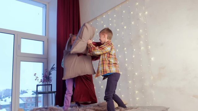 Children teen boy and girl playing fighting pillows together in bedroom with big window on Christmas holidays. Childhood, friend, friendship, fun concept. New Year interior with garland on wall, xmas.