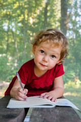 Little girl draws in a notebook on a background of nature. The beautiful, emotional face of a child of three years.