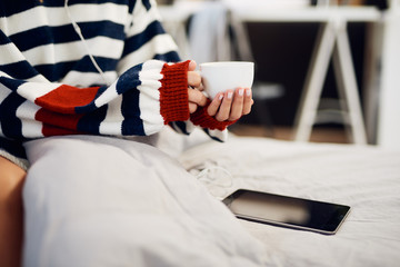 Caucasian woman in striped sweater sitting on bed, listening music over tablet and holding her fresh morning coffee.