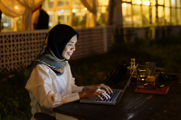 Smiling business asian muslim woman browsing on laptop at outdoor cafe in evening.