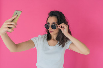 Beautiful girl in round sunglasses takes a selfie on the phone on a hot pink background