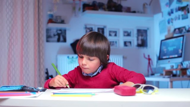 child sitting at a table draws with a pencil, looking at the camera
