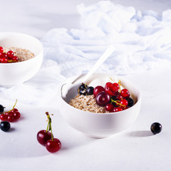 Raw oatmeal with yogurt and berries of cherry, currant and blueberry for making healthy breakfast in white ceramic bowl on a light background. Selective focus.