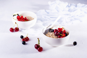 Raw oatmeal with yogurt and berries of cherry, currant and blueberry for making healthy breakfast in white ceramic bowl on a light background. Selective focus.