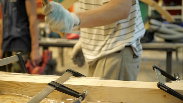 A craftsman in a carpentry workshop knocks with a rubber hammer on boards fastened with clamps