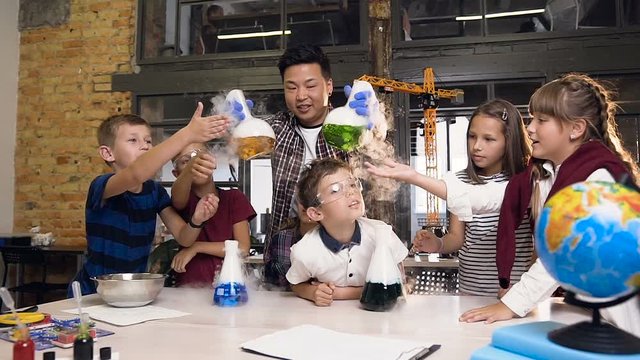 Front view. Asian male science teacher teaching young students doing chermical experiment, holding two flasks with liguid which causes intensive vaporization