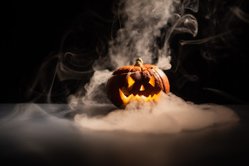 Halloween, orange pumpkin with a scary luminous face on a dark background. Gray thick smoke comes...