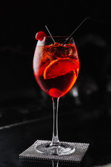An orange cocktail in a wine glass with an orange slice and a cherry. Aperol spritz. Dark background.