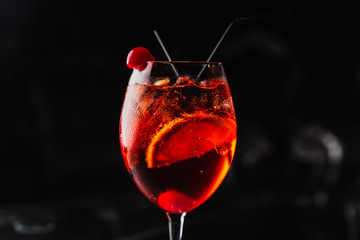 An orange cocktail in a wine glass with an orange slice and a cherry. Aperol spritz. Dark background.