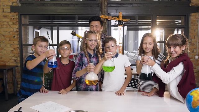Portrait shot of Korean male teacher with group of six cheerful young caucasian students wearing goggles during chemical experiment hold flaks with colored liguid where evaporates the cold smoke