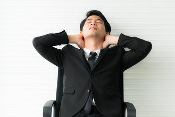 Young Asian handsome businessman wearing black suit arms behind head relaxing in his chair at office