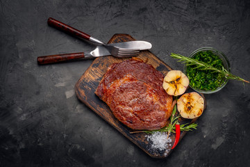 Grilled Chuck roll beef steak on a wooden board on dark stone background, near fork, knife and...