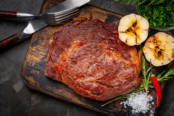 Well done grilled ribeye beef steak on a wooden board on dark stone background, near fork, knife and seasoning. Top view