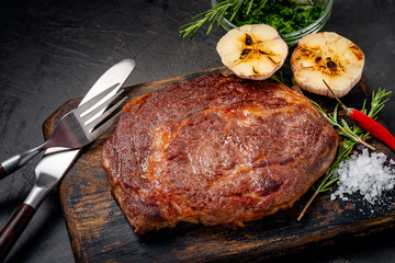 Well done grilled ribeye beef steak on a wooden board on dark stone background, near fork, knife...