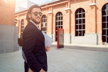 Photo of brunette in sunglasses and jacket with laptop standing in city on spring