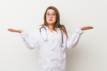Young doctor woman against a white wall confused and doubtful shrugging shoulders to hold a copy...
