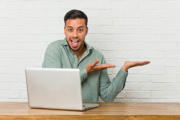 Young filipino man sitting working with his laptop excited holding a copy space on palm.