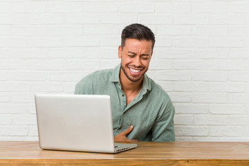 Young filipino man sitting working with his laptop laughs happily and has fun keeping hands on...