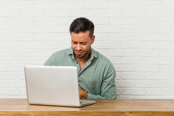 Young filipino man sitting working with his laptop sick, suffering from stomachache, painful disease concept.