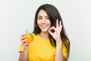 Young hispanic woman holding an orange cheerful and confident showing ok gesture.
