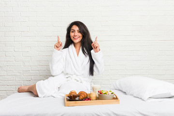 Young curvy woman taking a breakfast on the bed indicates with both fore fingers up showing a blank...