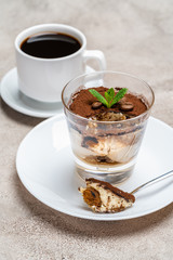 Portion of Classic tiramisu dessert in a glass and cup of coffee on concrete background