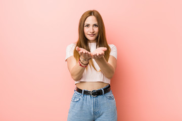 Young redhead ginger woman against a pink wall holding something with palms, offering to camera.