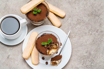 Portion of Classic tiramisu dessert in a glass, savoiardi cookies and cup of coffee on concrete background