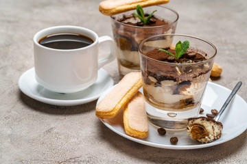 Portion of Classic tiramisu dessert in a glass, savoiardi cookies and cup of coffee on concrete background