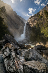 Wide angle view of the Yosemite Falls with rocks and boulders in the foreground, and a pine wood on the right