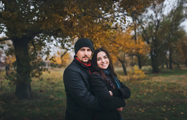 Young caucasian guy with mustache and beard and girl are standing embracing on the background of autumn nature. A pair of lovers dressed in warm clothes, a coat, a scarf, a hat.