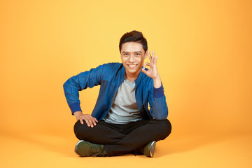 young casual asian man sitting with legs crossed and holding his head while smiling to the camera. isolated on color orange background