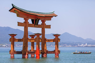A large wooden red torii emerging from the water with a traditional boat nearby and the distant skyline of Hiroshima in the background