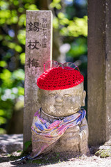 Close up of a small stone japanese jizo statue wearing a red wool cap against a green bokeh background