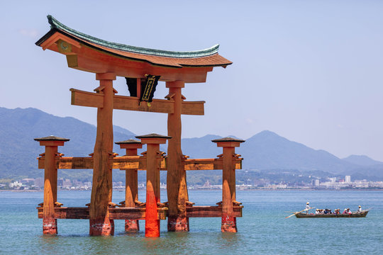 A large wooden red torii emerging from the water with a traditional boat nearby and the distant skyline of Hiroshima in the background