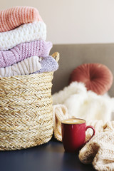 Bunch of stacked knitted pastel color sweaters with different knitting patterns perfectly folded in wicker basket on table, living room background. Fall winter season knitwear. Close up, copy space.