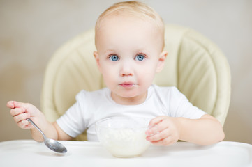 baby girl inquiringly looks at the camera, holding a spoon and about to eat porridge