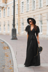 Fototapeta premium Elegant woman in long black dress, hat and sunglasses holding handbag standing and posing at city street. Stylish fashionable warm autumn look. Full length outdoor portrait